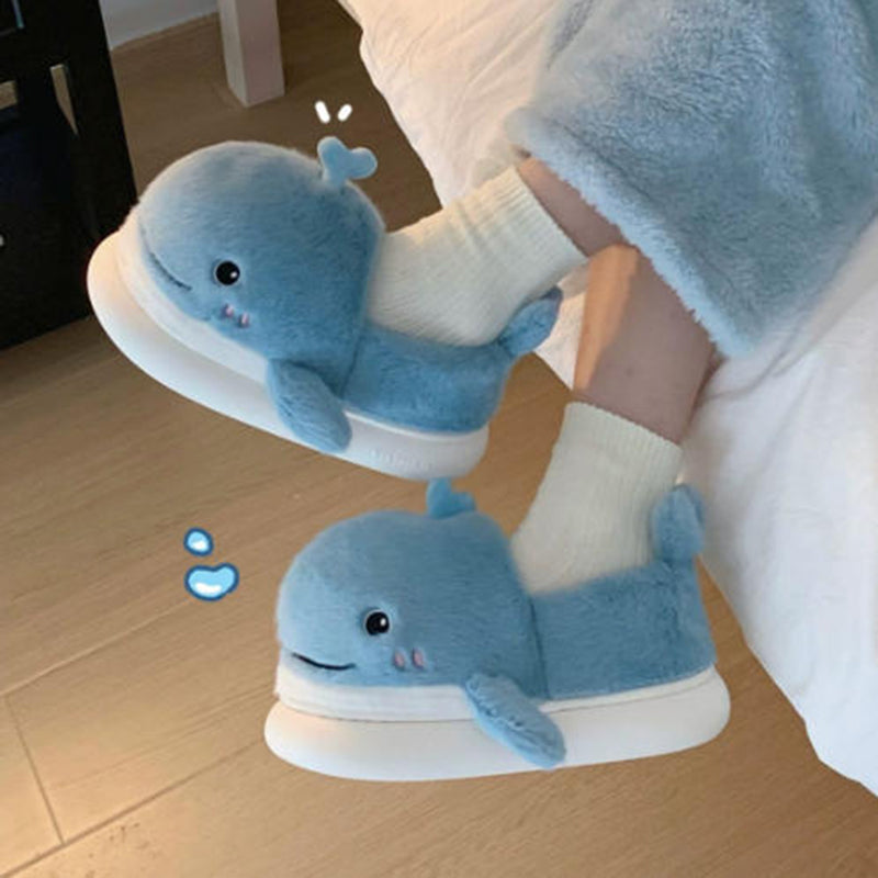 Light blue closed-back plush slippers shaped like a 3D whale with a spout and upturned tail, cozy indoor footwear with EVA sole
